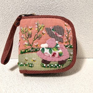 May include: A square fabric zipper pouch with a wrist strap, featuring an embroidered scene of a figure in a hat and floral dress standing in a garden with bees, flowers, and greenery. The pouch has a dark red and white checkered border.