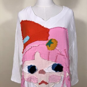 May include: White long-sleeve tunic with a V-neck and a large, colorful applique of a cartoon character's face. The applique features pink, orange, blue, and yellow patches, creating a whimsical design. The tunic is made of a textured fabric.