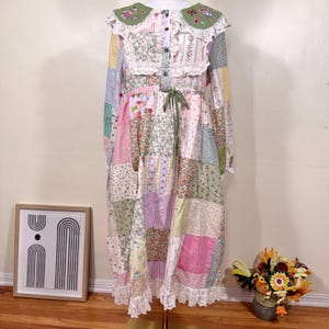 Puede incluir: Un vestido largo patchwork con cuello y puños de encaje blanco. El vestido presenta una variedad de telas estampadas florales y estampadas en rosa, verde y amarillo. Un cinturón de cinta verde ciñe la cintura. El cuello está adornado con detalles bordados.