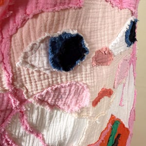 May include: Close-up of a textile art piece with a face-like design. The artwork features a light pink background with patches of white, pink, and orange fabric. The eyes are made of blue and black fabric, and the edges are frayed.