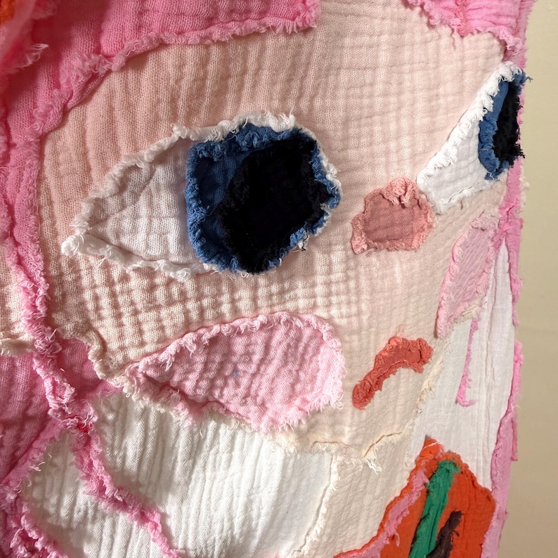 May include: Close-up of a textile art piece with a face-like design. The artwork features a light pink background with patches of white, pink, and orange fabric. The eyes are made of blue and black fabric, and the edges are frayed.
