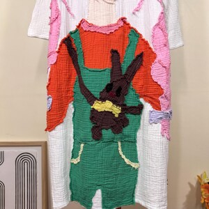 May include: White short-sleeved dress with a colorful patchwork design. The dress features an orange top, green overalls, and a brown bunny applique with a yellow bow. Pink and white accents adorn the sleeves and sides, creating a whimsical look.