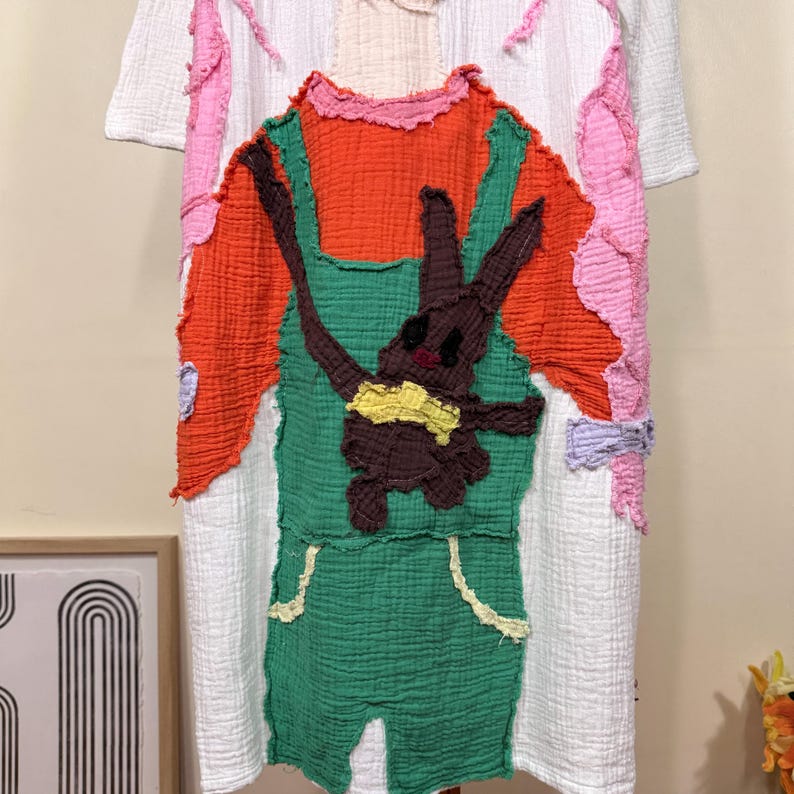 May include: White short-sleeved dress with a colorful patchwork design. The dress features an orange top, green overalls, and a brown bunny applique with a yellow bow. Pink and white accents adorn the sleeves and sides, creating a whimsical look.