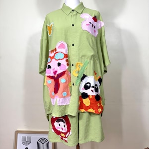 Puede incluir: Conjunto de camisa y pantalón corto verde claro. La camisa y el pantalón corto están adornados con coloridas y caprichosas aplicaciones de animales y formas de dibujos animados. El conjunto se exhibe en un maniquí.