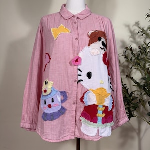 May include: A long-sleeved button-down shirt in a light pink gingham pattern. The front features large, colorful fabric appliqués including a white cartoon cat character, a purple and blue teacup, and a small yellow star shape.