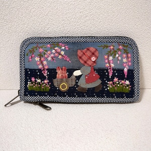 Pode incluir: Carteira de tecido com um design bordado. A frente apresenta uma figura com um chapéu vermelho puxando um carrinho com um coelho, sob ramos floridos. A borda é um padrão xadrez azul e branco.