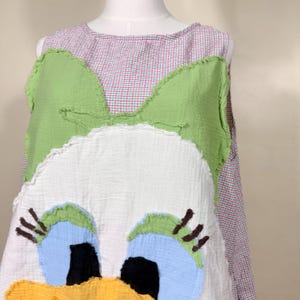 May include: A sleeveless dress featuring a whimsical duck design. The dress has a white base with a large duck face applique, including blue eyes, a yellow beak, and brown eyelashes. The top is a pink and white gingham pattern, with green accents.