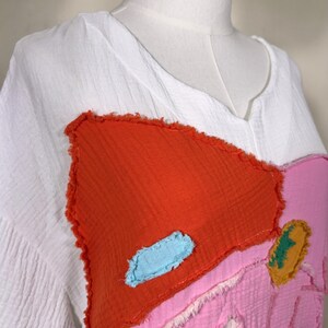 May include: White, textured fabric top with a V-neck and colorful patchwork design. The patchwork includes a large orange shape, a pink shape, and a small blue accent. The top is displayed on a mannequin.