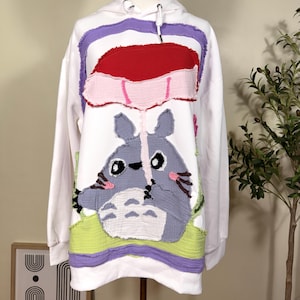 May include: A white hooded sweatshirt featuring a large, textured fabric appliqué of a grey Totoro character holding a red umbrella. The design includes frayed edges, light green accents, and purple trim, creating a layered, tactile appearance.