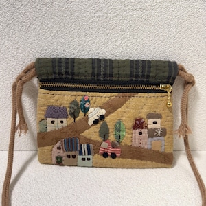 Puede incluir: Un pequeño bolso cruzado beige con una escena de pueblo caprichosa aplicada en la parte delantera. La escena incluye casas, árboles y coches. La solapa superior es de cuadros verdes y el bolso tiene una cremallera dorada y una correa de cuerda marrón.