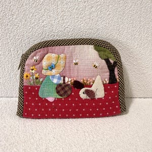 May include: A fabric zipper pouch featuring a quilted scene of a person in a yellow hat and green dress with a dog, flowers, and bees. The pouch has a red base with white dots, a pink checkered background, and a black and white checkered trim.