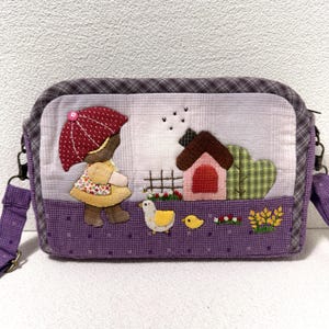 Puede incluir: Bolso bandolera acolchado hecho a mano con un diseño caprichoso. El bolso presenta una escena con una figura sosteniendo un paraguas rojo, una casa y un pato. El bolso tiene una correa morada y un patrón a cuadros en la parte superior e inferior.