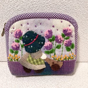 Könnte beinhalten: Handgefertigte, gesteppte Tasche mit einer skurrilen Gartenszene. Das Design zeigt eine Figur mit Hut, umgeben von gestickten lila und rosa Blumen. Die Tasche hat einen lila Vichy-Rand und einen Reißverschluss.