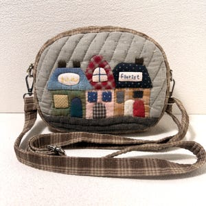 May include: A quilted crossbody bag with a charming village scene. The bag features a gray background with a stitched pattern, and a strap with a brown plaid design. The village scene includes houses with the words "Bakery" and "Florist".