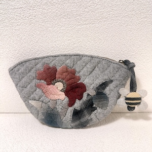 May include: A grey quilted pouch with a floral applique in shades of red, pink, and cream. The pouch has a zip closure and a bee charm attached to the pull. The pouch is approximately 15 cm wide.