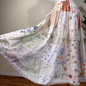 May include: A tiered maxi skirt featuring a patchwork design of various light-colored fabrics with floral, heart, and cartoon prints. The skirt has an elastic waistband and is accented with white lace trim along the horizontal seams and hemline.