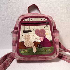 May include: A pink plaid quilted backpack featuring a front pocket with an appliqued scene of a person in a hat sitting next to a cat. The scene includes a brown fence, green grass with small flowers, musical notes, and a dark swing above.
