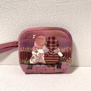 Puede incluir: Un pequeño estuche de tela rectangular en tonos rosa y morado, con un diseño de aplique de dos figuras sentadas en un banco. Las figuras están confeccionadas con diversos tejidos estampados, incluyendo telas florales y de cuadros, con una nota musical.