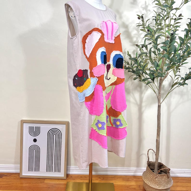 May include: A sleeveless beige dress featuring a large, colorful applique of a cartoon bear holding a cupcake. The bear has brown fur, blue eyes, and a pink chef's hat. The dress has a relaxed fit and a round neckline.