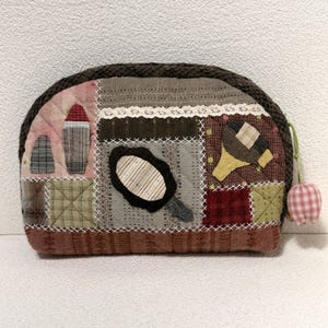 Puede incluir: Un neceser acolchado hecho a mano con cremallera marrón y una pequeña borla decorativa a cuadros rosas y blancos. La bolsa presenta un diseño de patchwork con varias telas en tonos marrones, rosas y verdes, y un pequeño espejo aplicado.