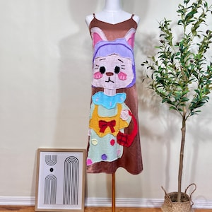 以下が含まれることがあります： 茶色のサテンのスリップドレス。気まぐれでカラフルなアップリケのデザイン。 アップリケは、ウサギのような顔をした漫画のキャラクターで、紫色の帽子、青い襟、赤いリボンが付いた黄色のエプロンを着ています。赤いハンドバッグがドレスの右側にあります。
