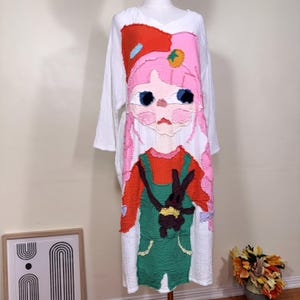 May include: White long-sleeve dress featuring a colorful cartoon character design. The design includes a pink-haired girl wearing a red hat and green overalls, holding a brown bunny. The dress has a V-neck and a loose fit.