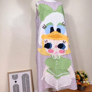 May include: A sleeveless, lilac-checkered dress featuring a large, whimsical appliqu&eacute; of a cartoon character. The character has a white face, blue eyes, and a yellow beak and hair. The dress is displayed on a mannequin.