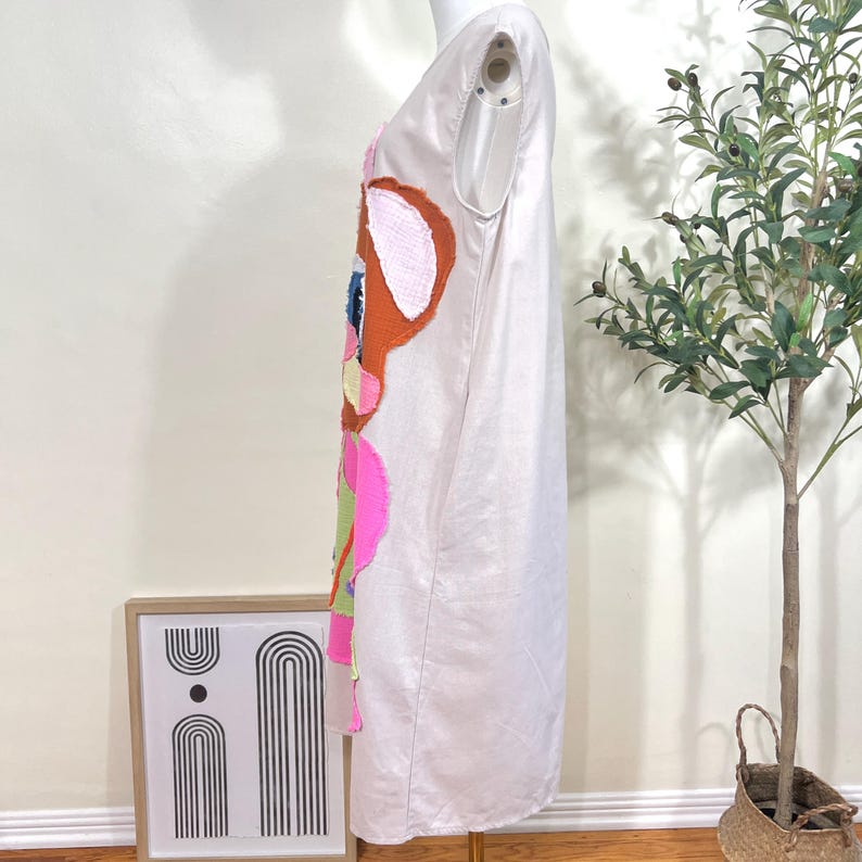 May include: A light beige sleeveless dress featuring a colorful cartoon deer applique on the front. The deer design includes shades of brown, pink, blue, and green. The dress is displayed on a mannequin.