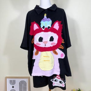 Könnte beinhalten: Schwarzes kurzärmeliges Hemd und Shorts-Set mit einem skurrilen, farbenfrohen katzenartigen Kreaturen-Design. Die Kreatur hat eine rote Kapuze, einen rosa Körper und einen Cupcake auf dem Kopf. Die Shorts haben ein passendes Design.