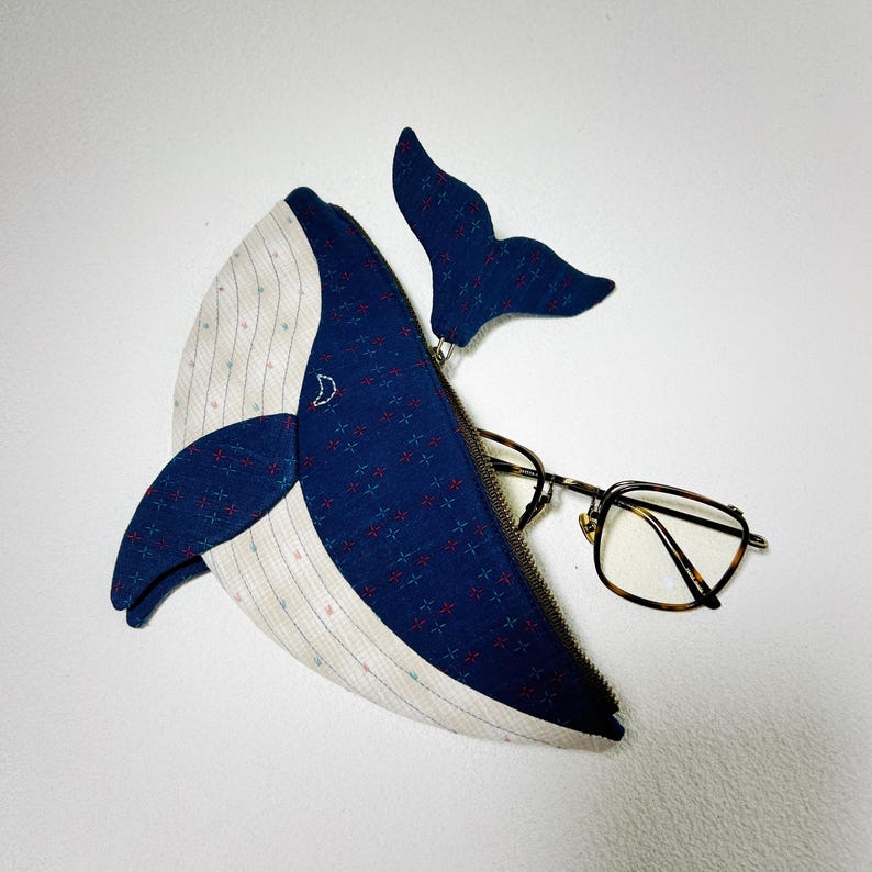 K&ouml;nnte beinhalten: Eine blaue, walf&ouml;rmige Rei&szlig;verschlusstasche aus einem Stoff mit wei&szlig;em und blauem Muster. Die Tasche ist offen und eine Brille liegt darin.