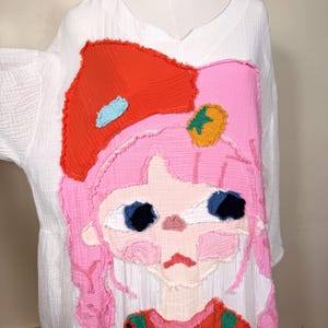 May include: White, loose-fitting dress with a large, colorful cartoon graphic on the front. The graphic features a pink-haired girl with a red bow, blue eyes, and a red and green outfit. The dress has a V-neck and short sleeves.