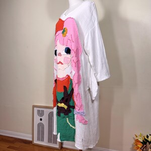May include: White, long-sleeved dress with a colorful, textured appliqu&eacute; of a cartoon girl. The girl has pink hair, a red hat, and a green and red outfit. The dress has a V-neck and a loose fit.