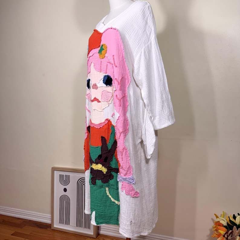May include: White, long-sleeved dress with a colorful, textured appliqu&eacute; of a cartoon girl. The girl has pink hair, a red hat, and a green and red outfit. The dress has a V-neck and a loose fit.