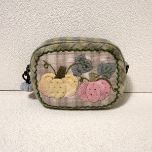 May include: A small, rectangular fabric pouch featuring a quilted beige and green plaid pattern. The front panel displays an appliqued design of two pumpkins, one yellow and one pink, with green fabric leaves and vines, finished with green stitching.