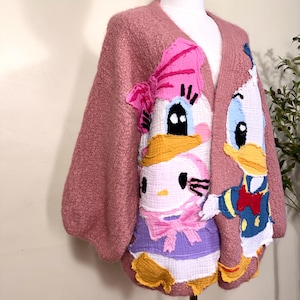 Peut inclure: Un gilet ouvert rose poudré orné de grands appliqués en tissu à bords francs représentant des personnages de dessins animés, dont un chat blanc avec un nœud rose, une cane avec un nœud rose et un canard en tenue de marin bleu avec un nœud papillon rouge.