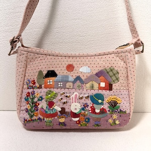 May include: A light pink quilted shoulder bag featuring a fabric appliqué scene of a village with houses, a sun, and a cloud. The foreground shows two figures and a white rabbit surrounded by colorful flowers, green foliage, and small bees.
