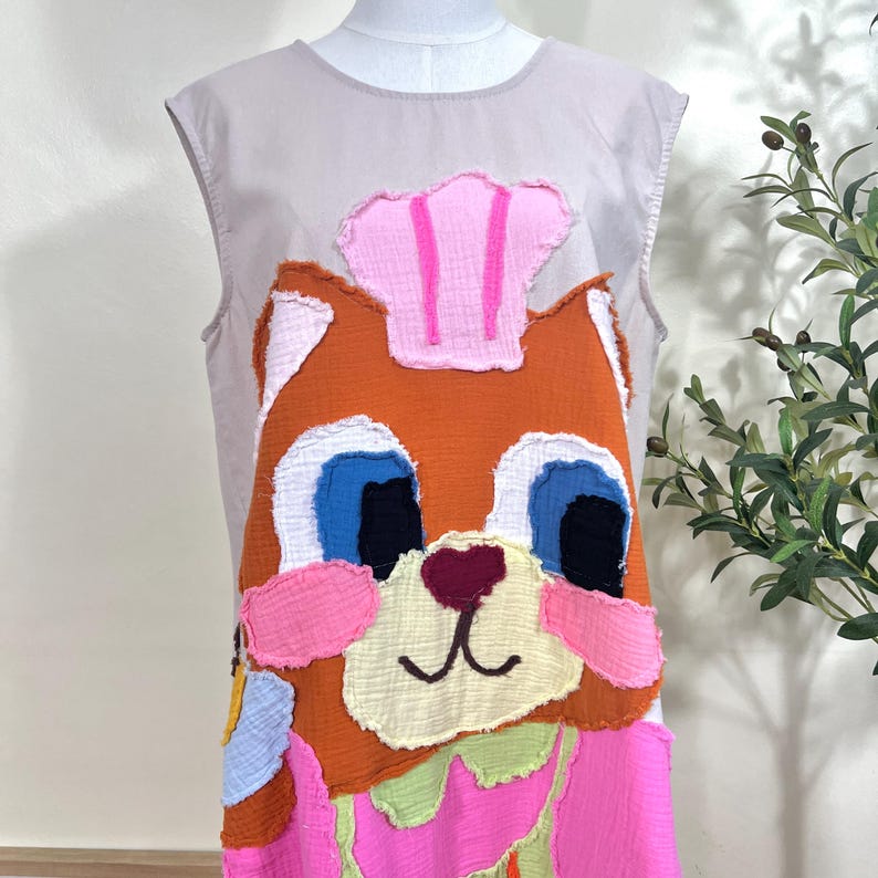 May include: Sleeveless dress featuring a whimsical cat design. The cat's face is rendered in orange, white, blue, and pink fabric, with a pink chef's hat. The dress has a light gray bodice and a colorful, patchwork-style design.