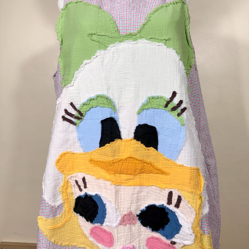 May include: A sleeveless dress featuring a whimsical duck face design. The dress has a white base with a large duck face applique in yellow, blue, black, and green. The dress is trimmed with a pink and white gingham pattern.