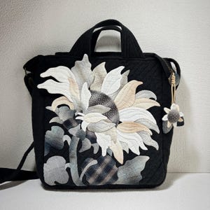 May include: A black quilted tote bag with a large sunflower design made from patchwork fabric. The sunflower is in shades of white, gray, and beige. The bag has a top zipper closure and a detachable strap.