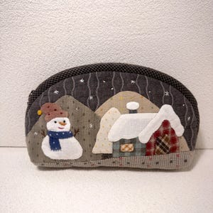 May include: A fabric coin purse depicting a winter scene. It features a snowman wearing a blue scarf and a house with a snow-covered roof. The purse has a dark grey zip and a patterned border.