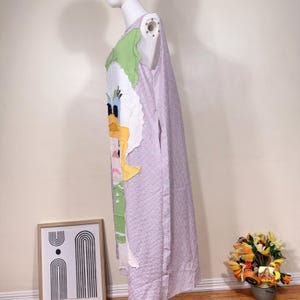 May include: A sleeveless, knee-length dress featuring a cartoon duck design on the front. The dress has a light purple and white checkered pattern and is displayed on a mannequin. The duck design includes yellow, green, and white fabric.