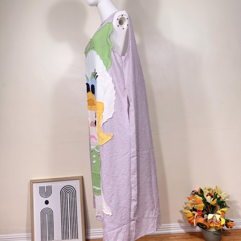 May include: A sleeveless, knee-length dress featuring a cartoon duck design on the front. The dress has a light purple and white checkered pattern and is displayed on a mannequin. The duck design includes yellow, green, and white fabric.