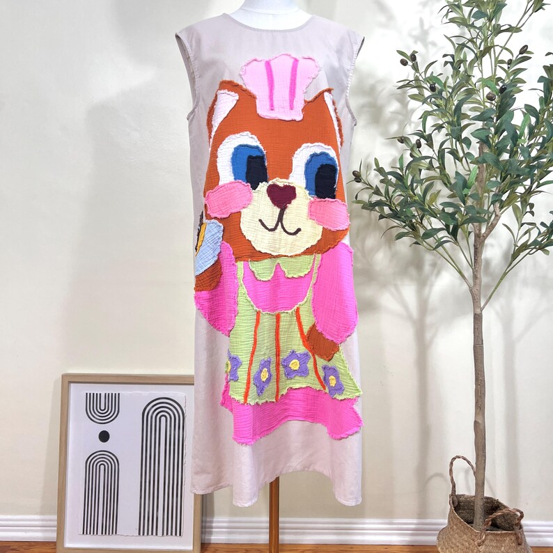 May include: A sleeveless, light beige dress featuring a large, colorful applique of a cartoon bear chef. The bear has brown fur, blue eyes, and a pink chef's hat and apron. The dress is displayed on a mannequin.