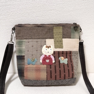 Puede incluir: Bolso bandolera patchwork con correa marrón ajustable. El bolso presenta una variedad de cuadrados de tela en tonos marrón, verde y beige. En la parte delantera hay un gato y dos tortugas aplicados. El bolso tiene cierre de cremallera.