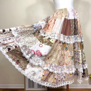 Pode incluir: Uma saia patchwork com um design em camadas. A saia apresenta vários padrões de tecido em tons de castanho, bege e branco, com estampas florais e geométricas. A renda branca adorna cada camada, criando uma estética boêmia fluida.