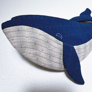 K&ouml;nnte beinhalten: Ein blaues und graues Pl&uuml;schtier in Form eines Wals mit einem Rei&szlig;verschluss. Der Wal ist aus Stoff mit einem Muster aus kleinen roten Kreuzen. Der Bauch des Wals ist aus einem helleren grauen Stoff mit einem Muster aus d&uuml;nnen, wellenf&ouml;rmigen Linien.