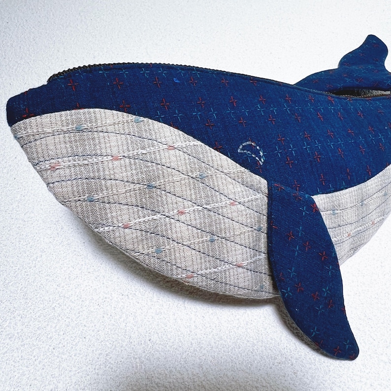 K&ouml;nnte beinhalten: Ein blaues und graues Pl&uuml;schtier in Form eines Wals mit einem Rei&szlig;verschluss. Der Wal ist aus Stoff mit einem Muster aus kleinen roten Kreuzen. Der Bauch des Wals ist aus einem helleren grauen Stoff mit einem Muster aus d&uuml;nnen, wellenf&ouml;rmigen Linien.