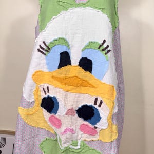 May include: A sleeveless dress featuring a whimsical cartoon character design. The dress has a white base with a large, appliqued face, including blue eyes, a yellow hat, and pink cheeks. The dress is trimmed with green and a red and white checkered pattern.