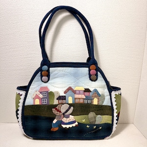 May include: A quilted tote bag featuring a fabric appliqué scene of a village with colourful houses and two figures sitting in a field. The bag has dark blue handles, white lace trim on the sides, and decorative fabric-covered buttons at the base.