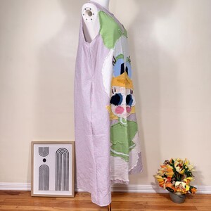 May include: A sleeveless, patchwork dress featuring a cartoon character design. The dress has a light purple and white checkered pattern on the left side, and a colorful cartoon face on the right side. The dress is displayed on a mannequin.